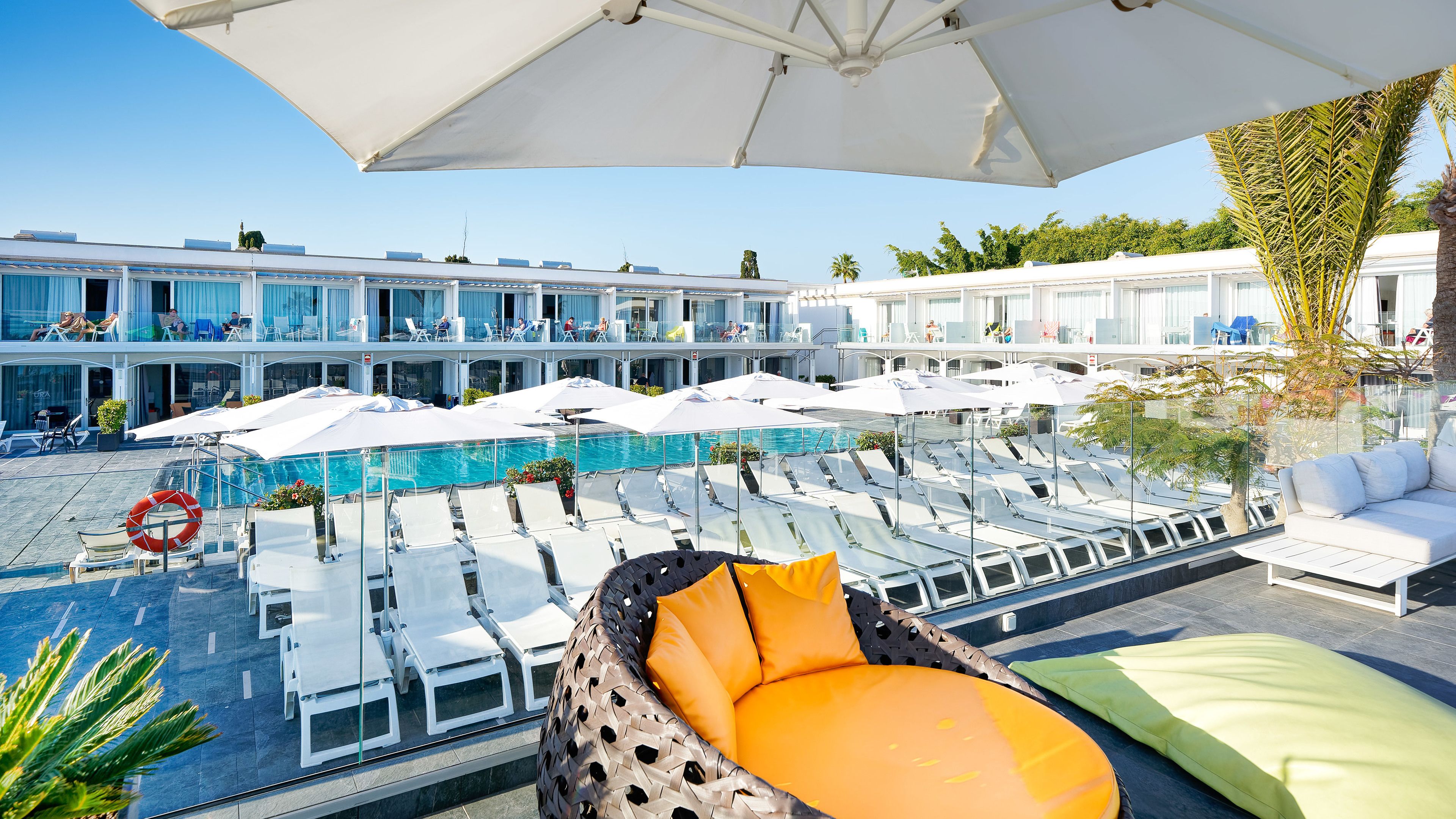 A rooftop pool area with lounge chairs and umbrellas, a clear view of the pool, and a building with multiple balconies in the background.
