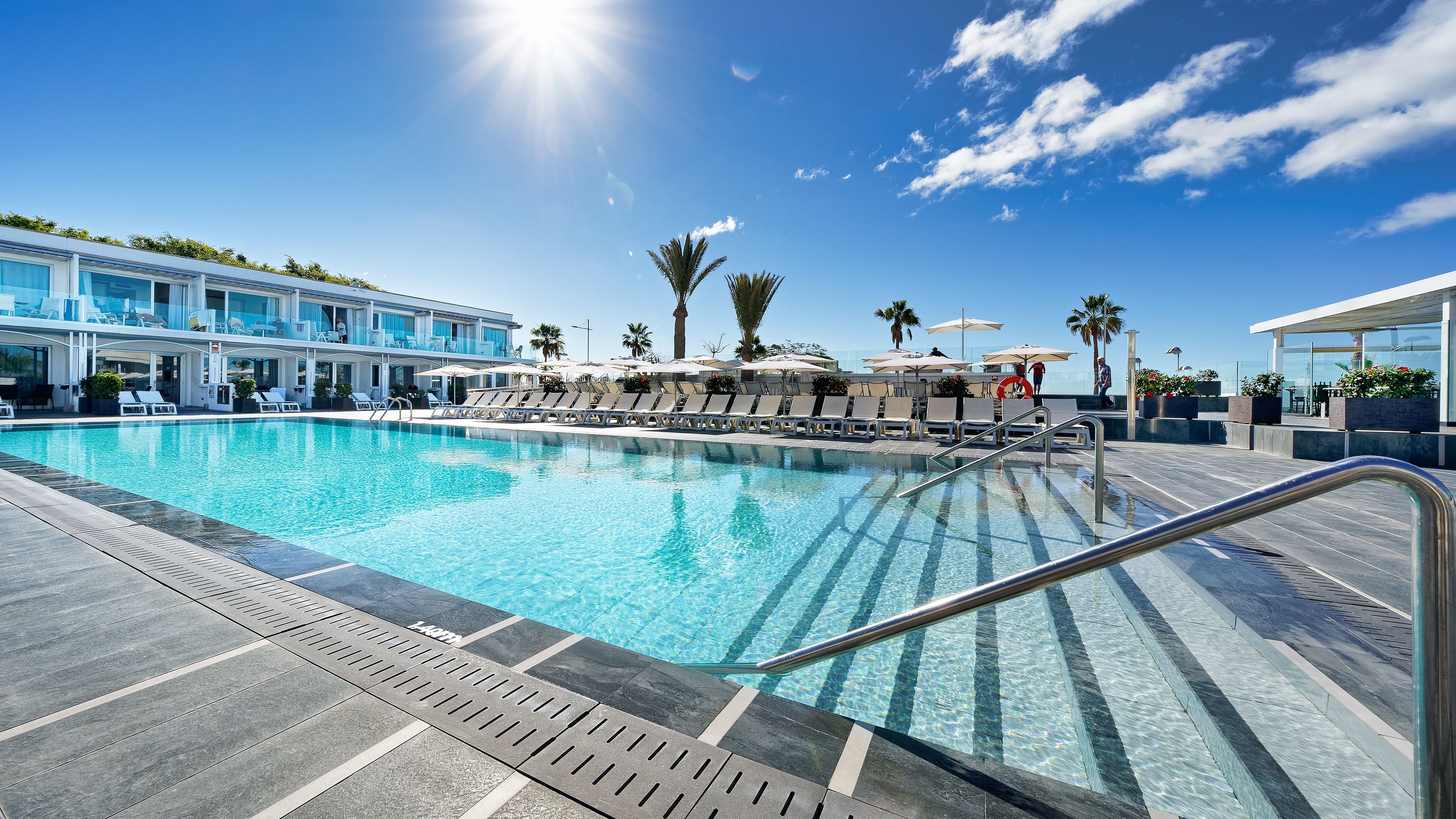 A swimming pool with lounge chairs and umbrellas, surrounded by a modern building with a glass facade.