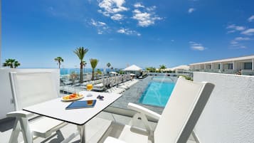 A rooftop pool area with lounge chairs, a dining table with food and drinks, and a clear view of the ocean.