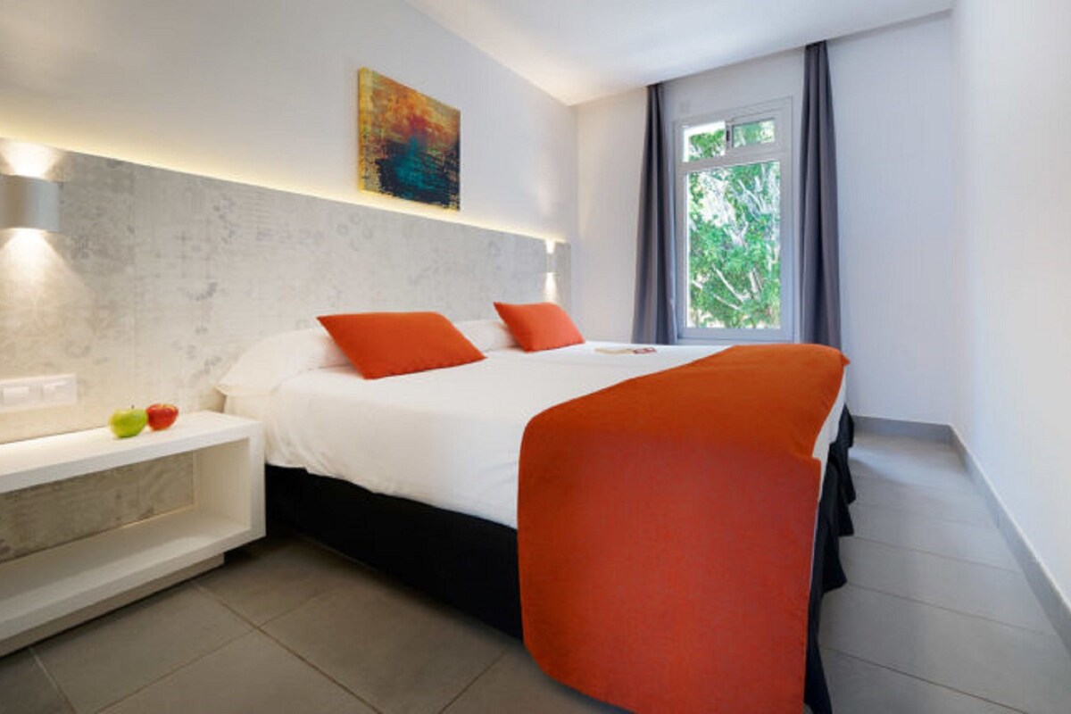 Habitación de hotel moderna con una cama grande, una mesita de noche con manzanas, un cuadro sobre la cama y una ventana con vistas a árboles.