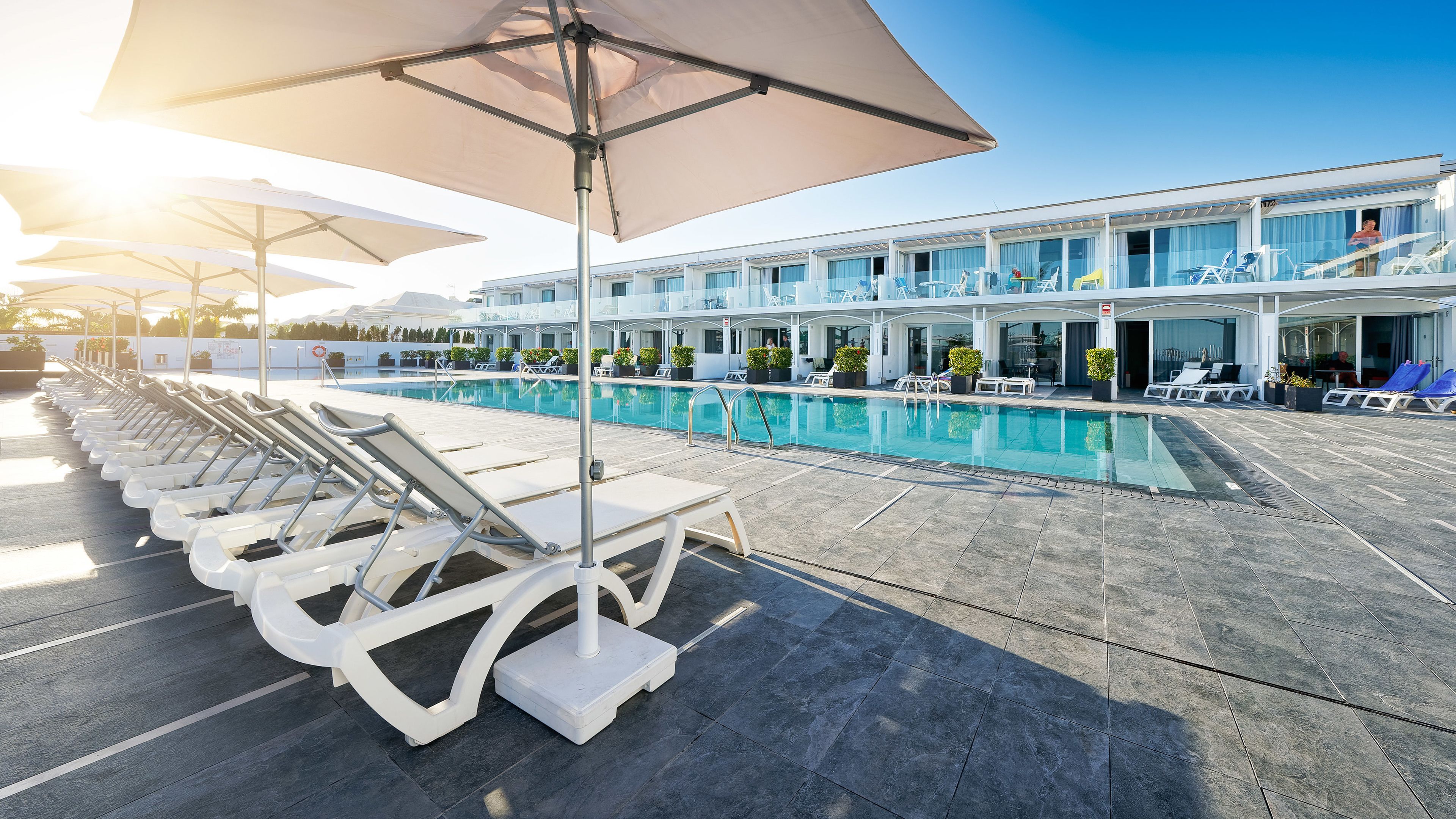 Un espace aménagé au bord de la piscine, avec des chaises longues, des parasols et un bâtiment doté de nombreuses fenêtres et de balcons.