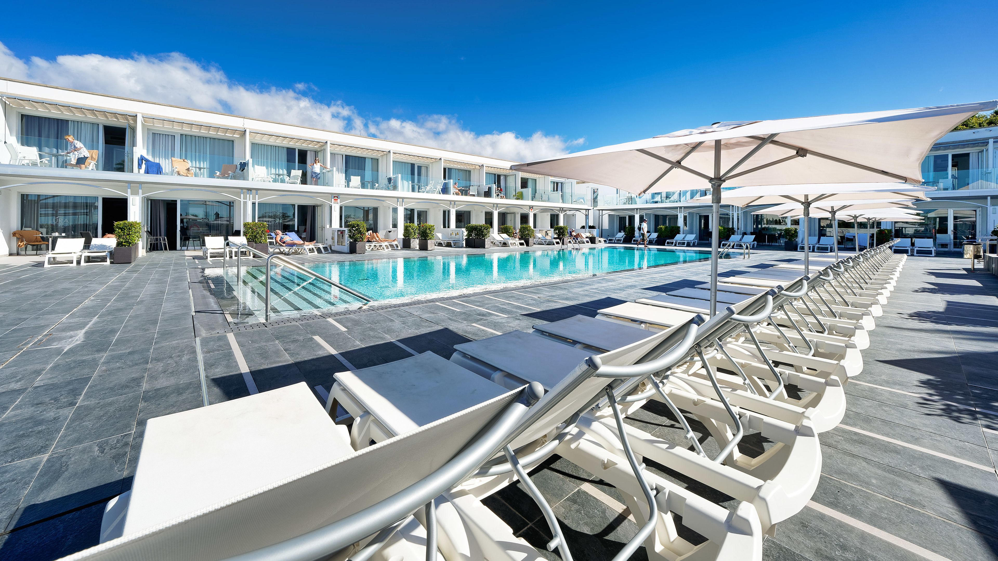 Area piscina con lettini e ombrelloni, una piscina e un edificio con numerosi balconi.