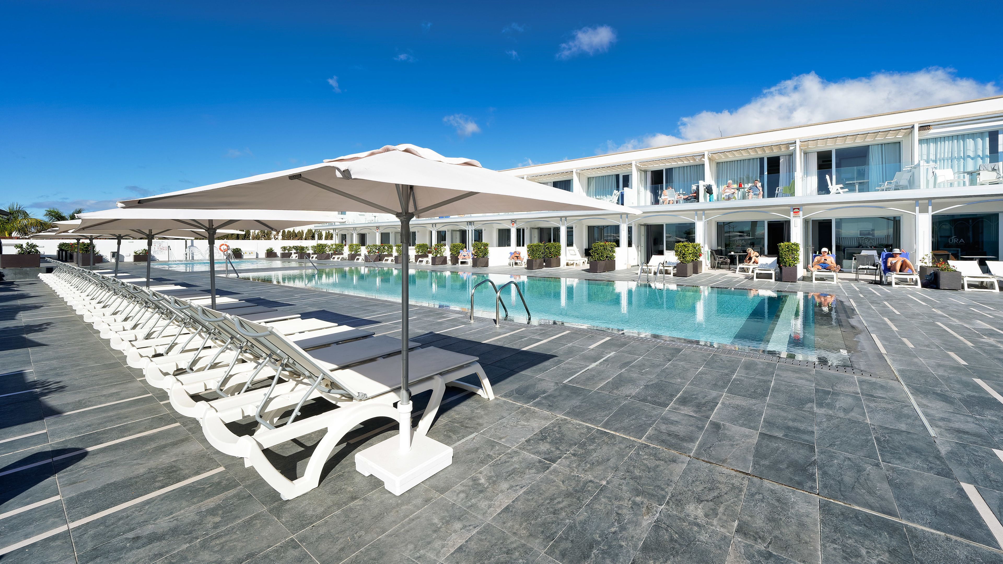 Zona de piscina con sillones de playa y sombrillas, cielo azul despejado y un edificio con ventanas amplias y un porche cubierto.
