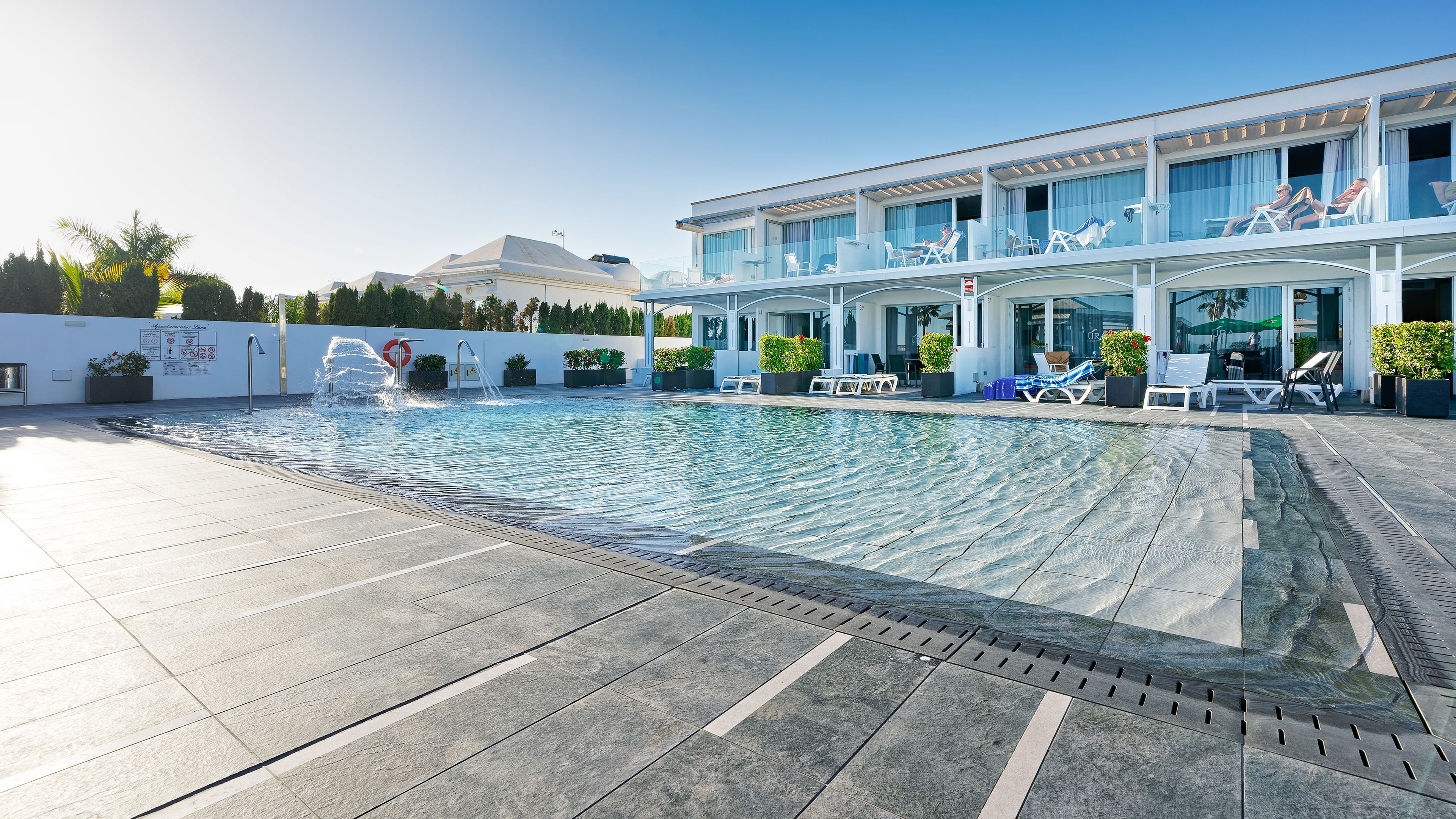 A swimming pool with a water feature, lounge chairs, and a building with large windows and a covered balcony.