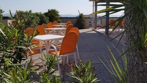 Un espace de restauration en plein air avec des tables blanches et des chaises orange, entouré de verdure et offrant une vue sur la mer.