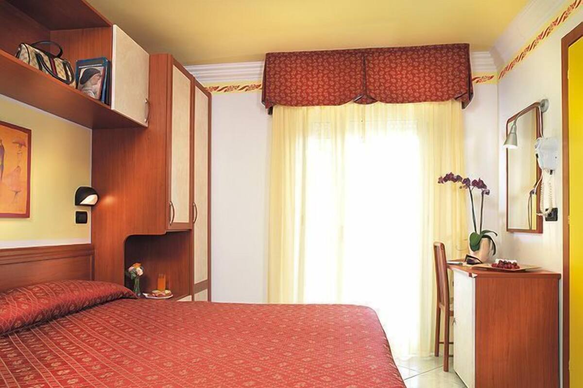 Ein Hotelzimmer mit einem großen Bett, einem Schreibtisch mit Stuhl, einem Spiegel und einem Regal mit Büchern und einer Handtasche.
