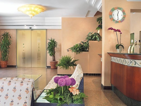 A hotel lobby with a reception desk, a sofa with a floral arrangement, and potted plants. - Hotel Grado (Bellaria-Igea Marina)