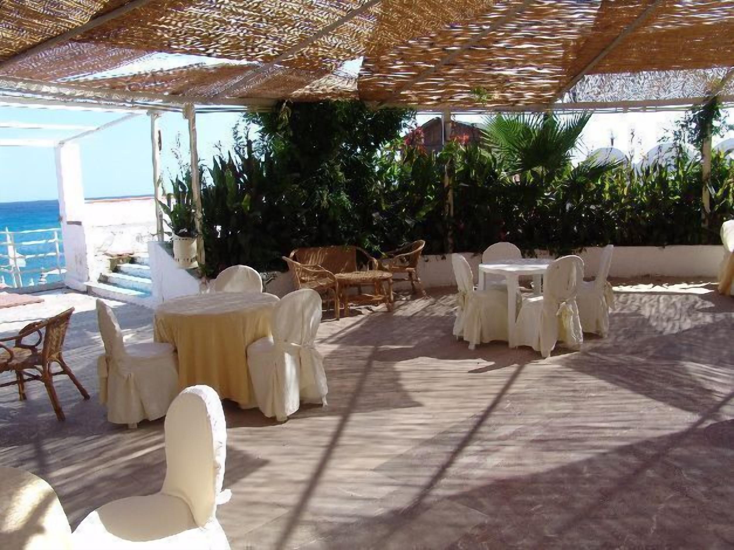 An outdoor patio area with round tables and chairs, covered by a woven canopy, overlooking a scenic ocean view.