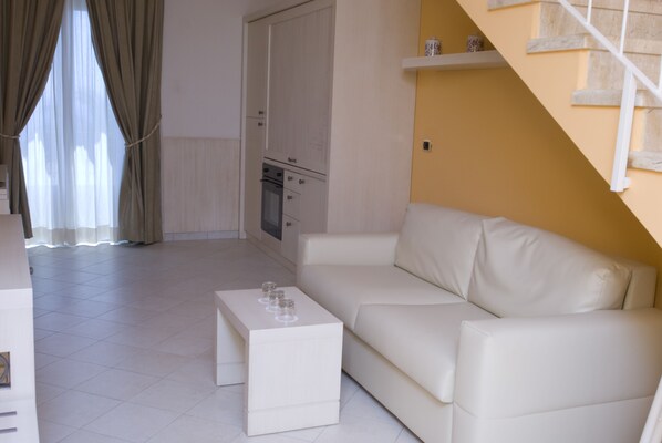 Una sala de estar con un sofá blanco, una pequeña mesa de centro blanca y una zona de cocina con estufa y armarios.