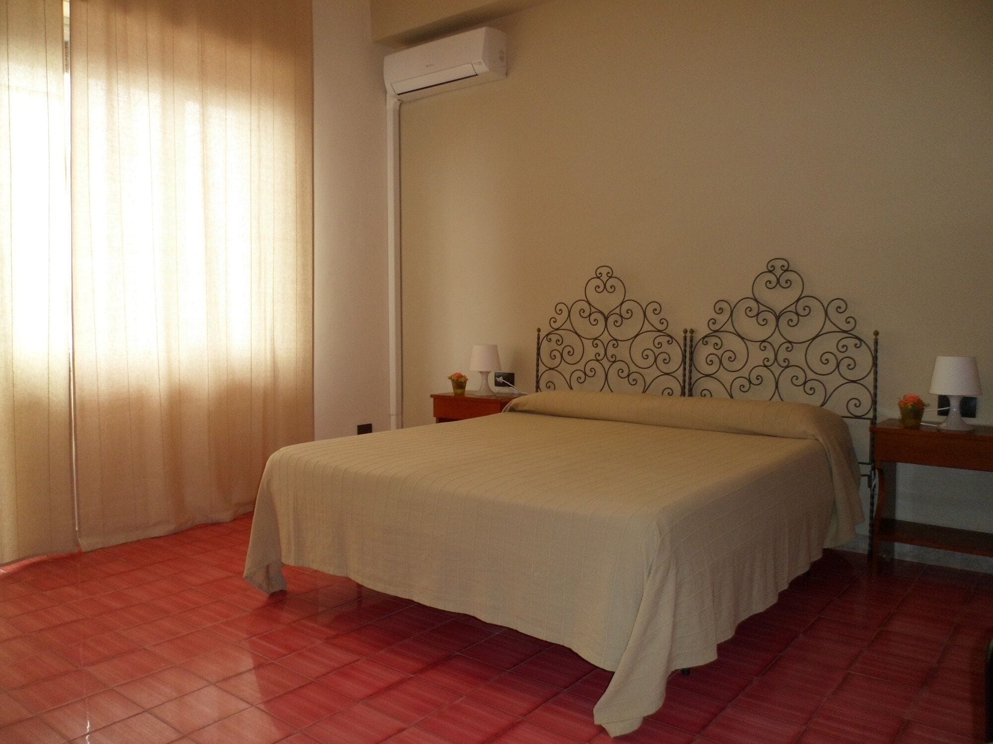A neatly made bed in a room with a red tiled floor, a white air conditioning unit, and a decorative metal headboard.