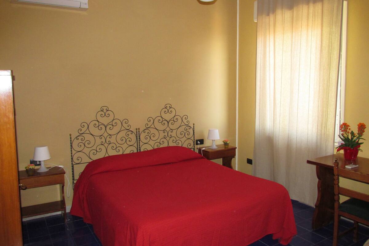 A room with a bed, a nightstand, a lamp, a table with a vase of flowers, and a window with curtains.