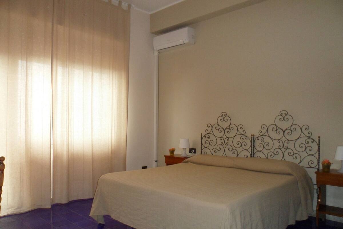 A bedroom with a bed, curtains, a nightstand, a lamp, and a wall-mounted air conditioner.