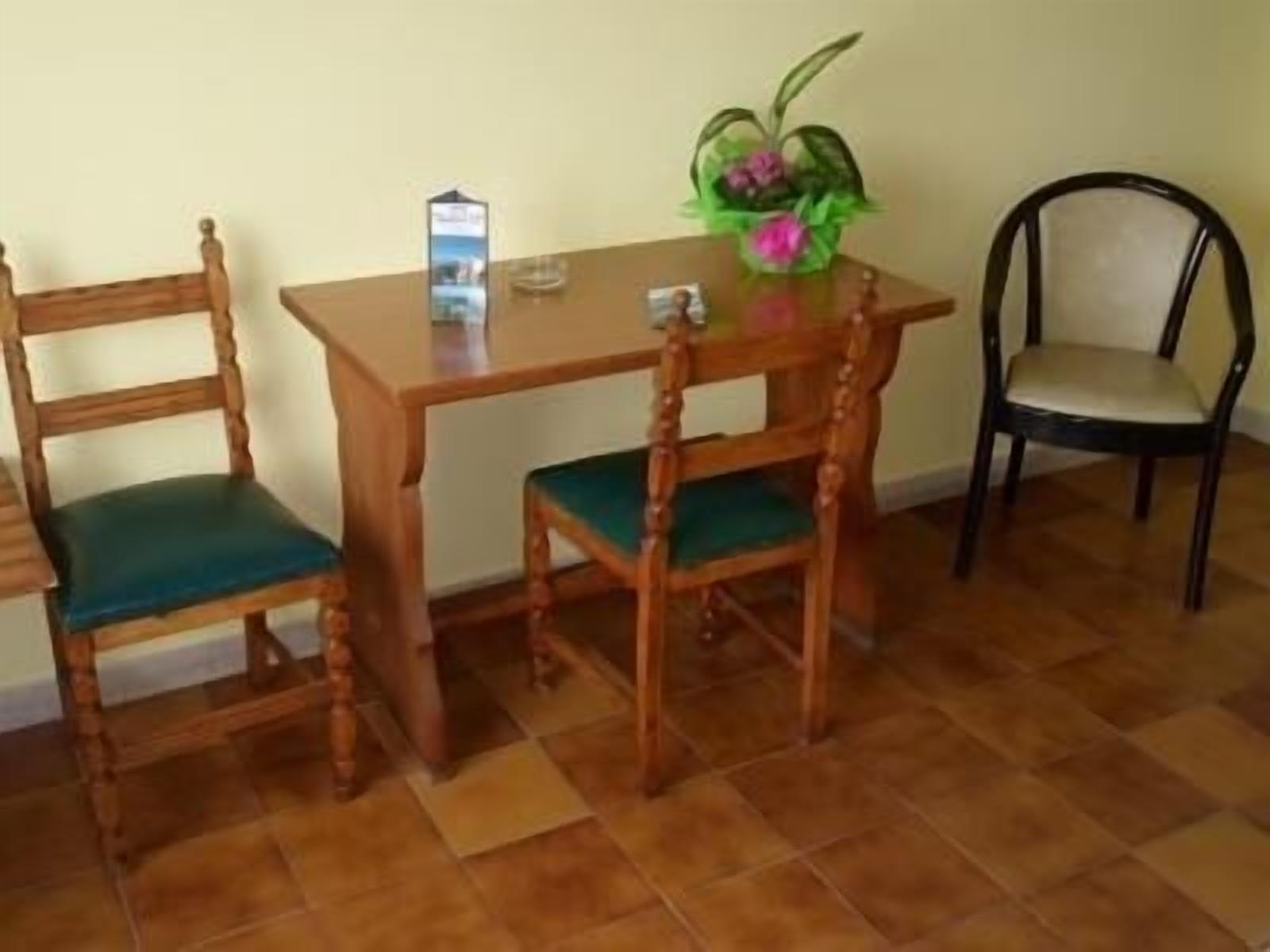 A wooden dining table with two chairs, one with a green cushion and another with a beige cushion. A small potted plant with pink flowers is on the table.