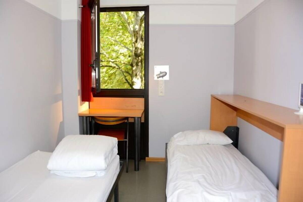 A small, compact room with two single beds, a desk, and a window with a view of trees.