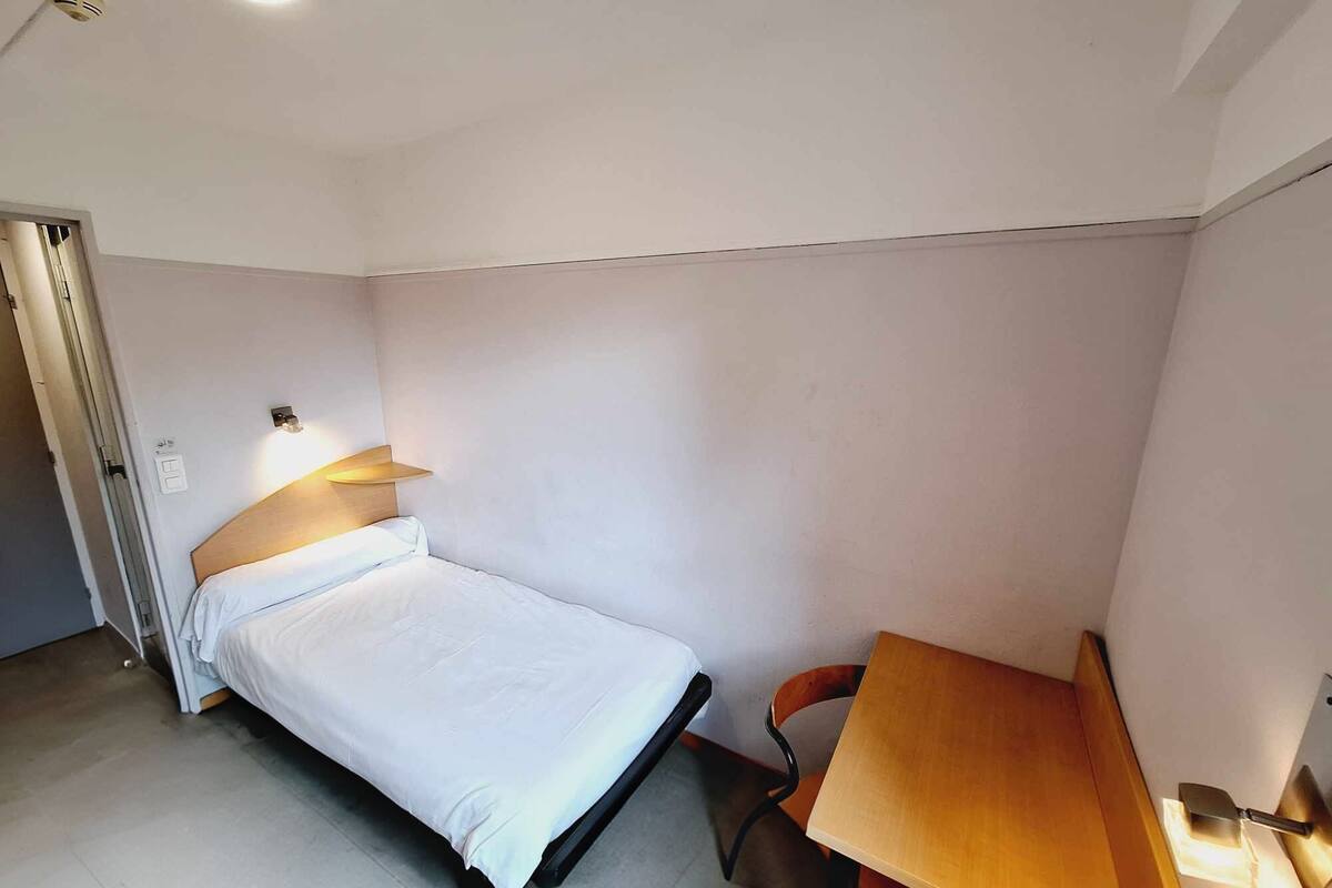 A small, clean room with a bed, a desk, and a chair. There is a ceiling light and a wall-mounted light above the bed.