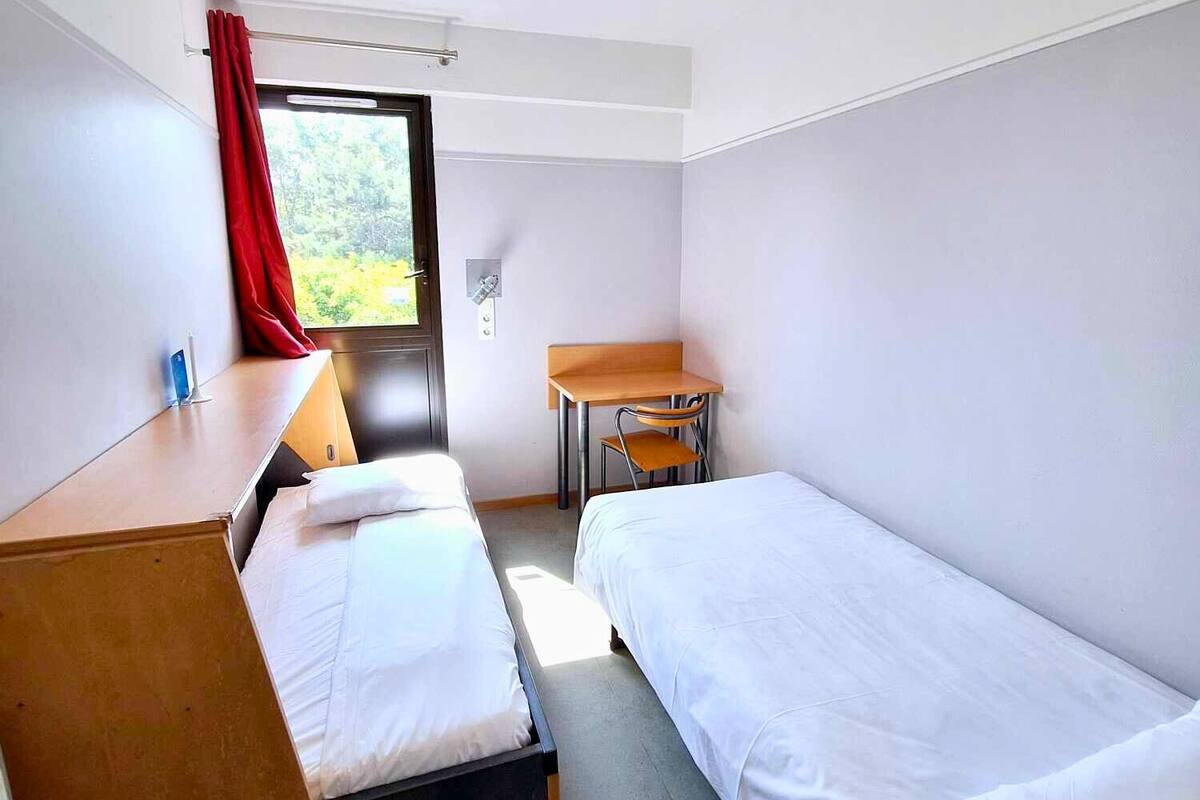 A hotel room with two single beds, a desk, a chair, and a window with a red curtain.