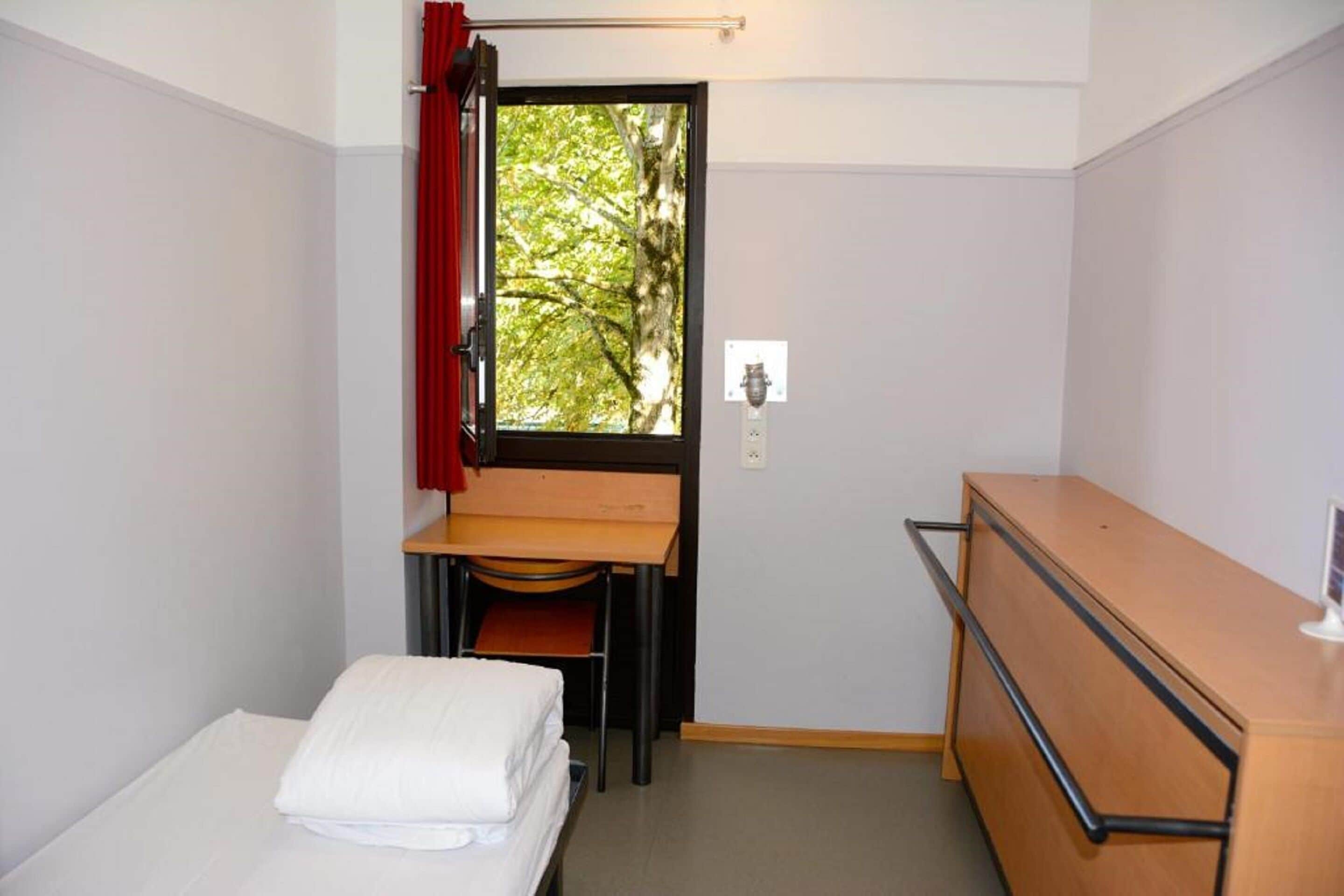 A compact room with a bed, a desk, and a window with a view of trees.