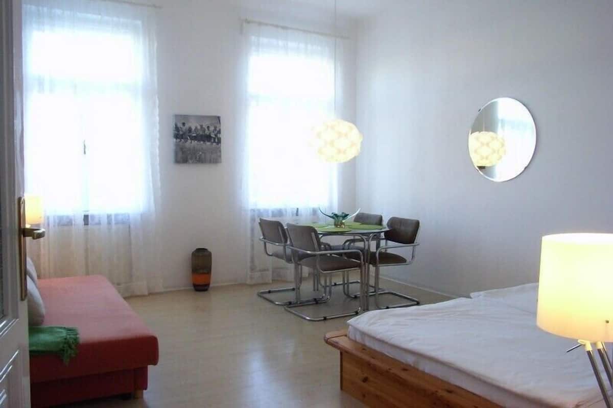 A room with a bed, a red sofa, a dining table with chairs, and two windows with curtains.