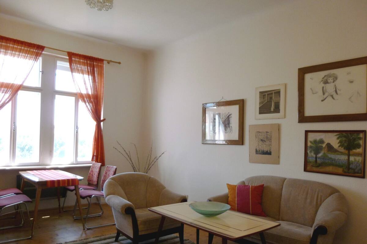 A living room with a sofa, armchairs, a coffee table, and a dining table. There are framed pictures on the wall and a window with curtains.