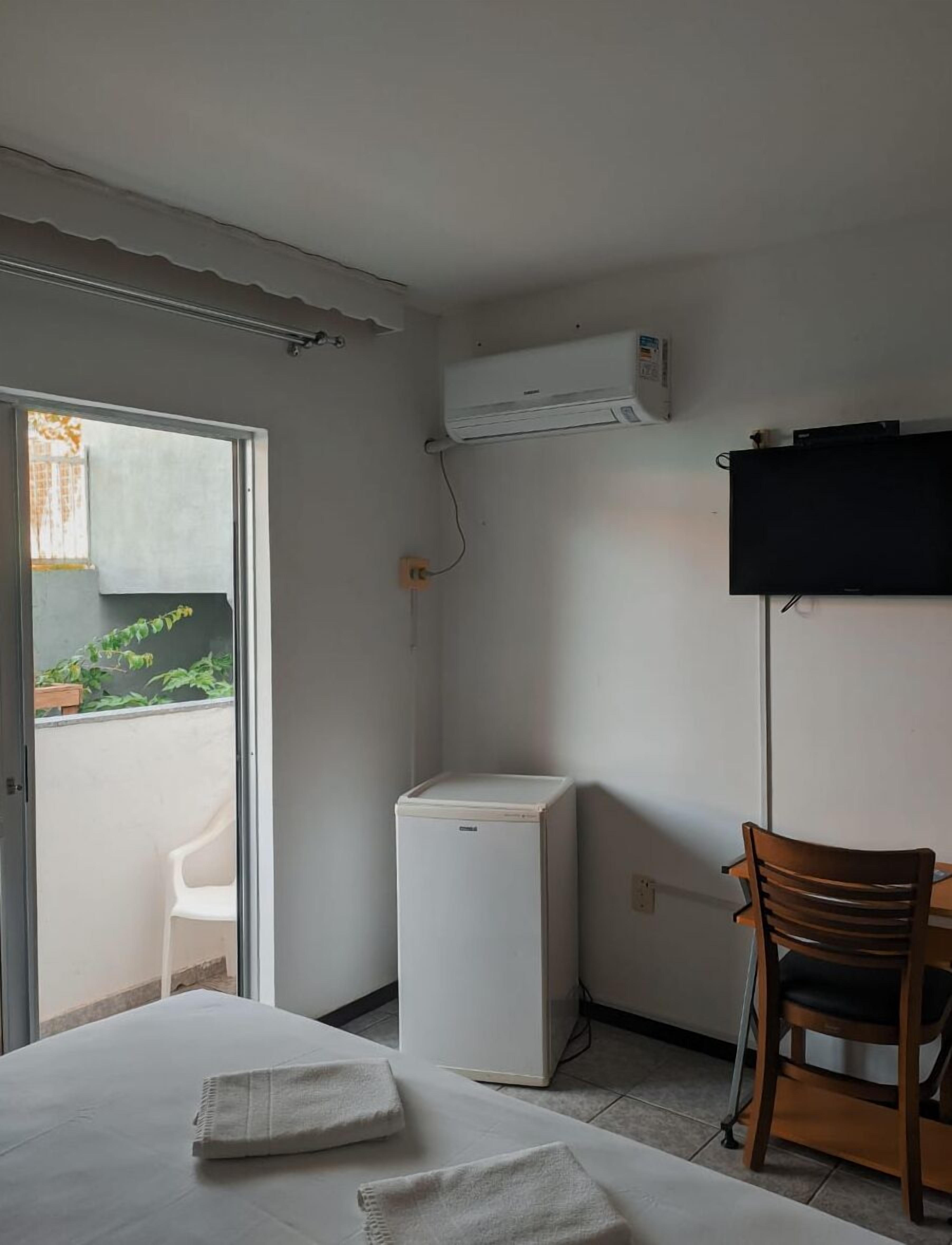 A room with a bed, a white air conditioner, a wooden chair, and a television mounted on the wall.