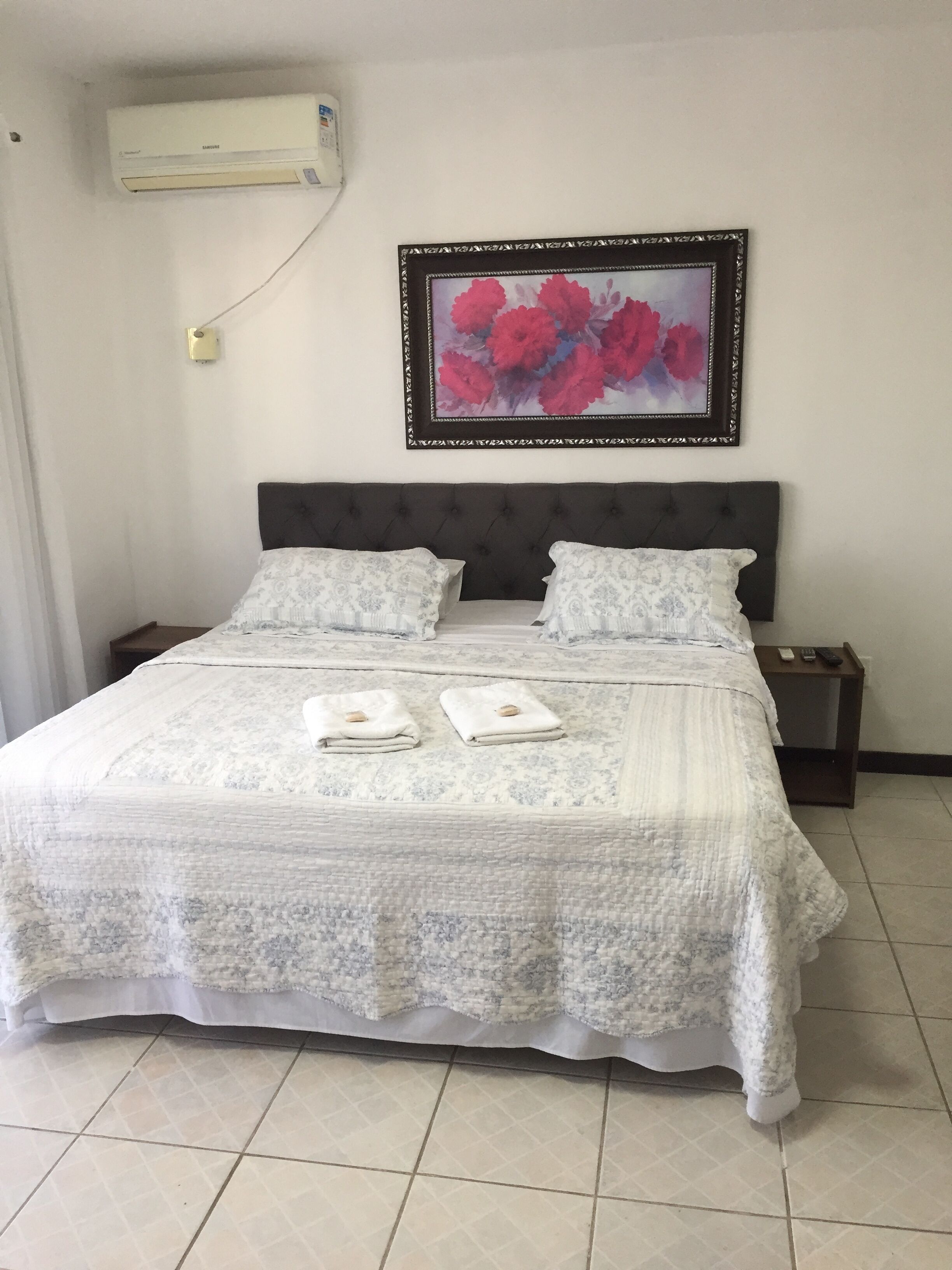 Un lit bien fait, recouvert d’une couvre-lit à motifs, avec deux serviettes à portée de main, une œuvre d’art florale encadrée au-dessus du lit, une table de chevet avec une télécommande et un appareil de climatisation fixé au mur.