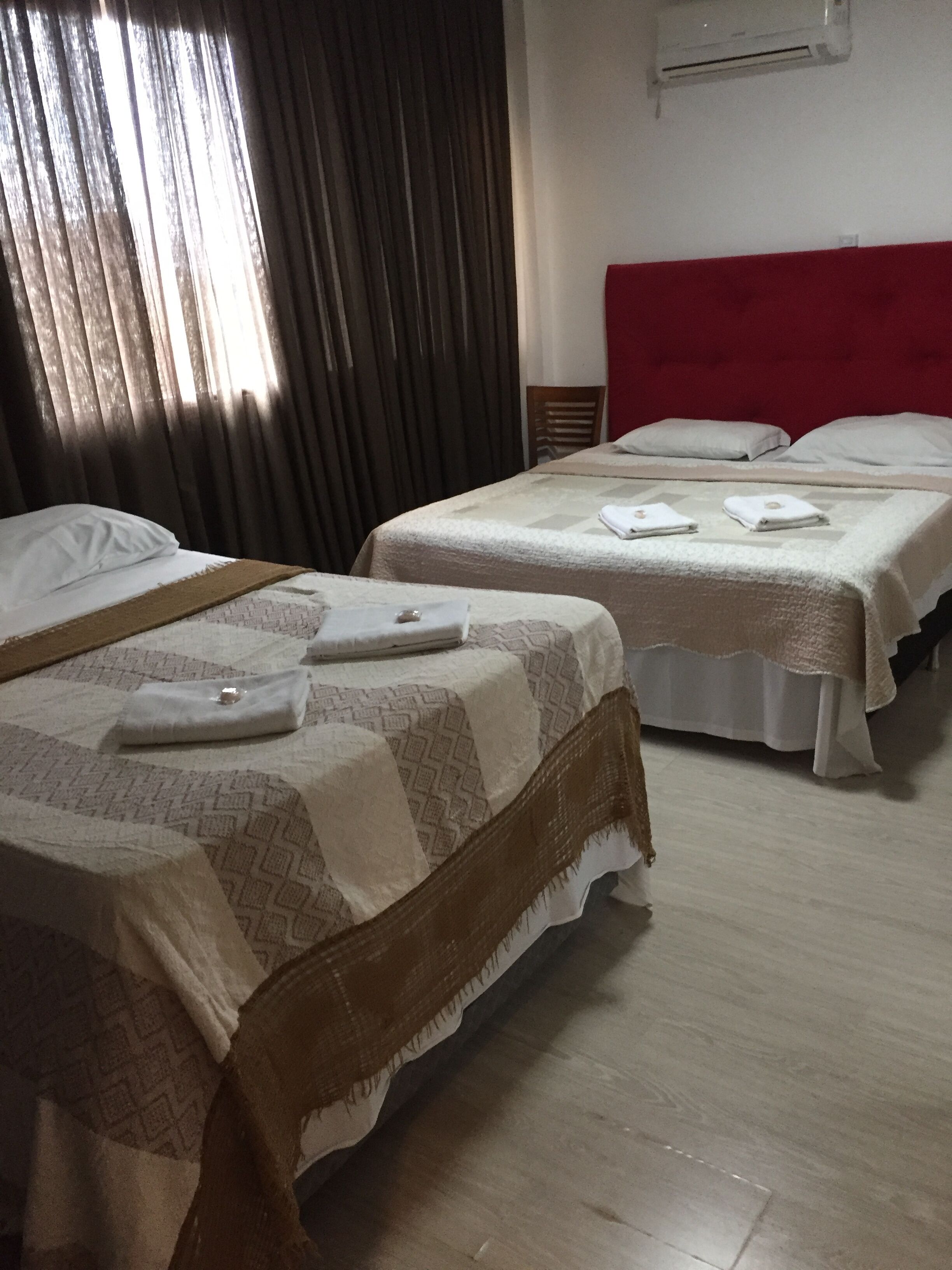 Quarto de hotel com duas camas, cada uma com colcha e almofadas estampadas. Uma cama tem cabeceira vermelha e a outra, de madeira. É visível uma janela com cortinas, e um aparelho de ar condicionado fixado na parede.