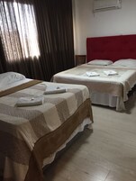 Quarto de hotel com duas camas, cada uma com colcha e almofadas estampadas. Uma cama tem cabeceira vermelha e a outra, de madeira. É visível uma janela com cortinas, e um aparelho de ar condicionado fixado na parede.