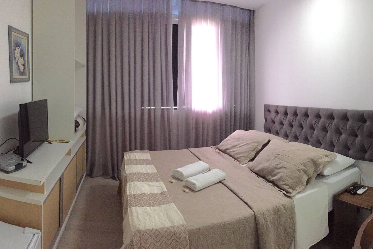 A hotel room with a bed, a desk with a computer, a TV, a window with curtains, and a bathroom visible through a glass door.