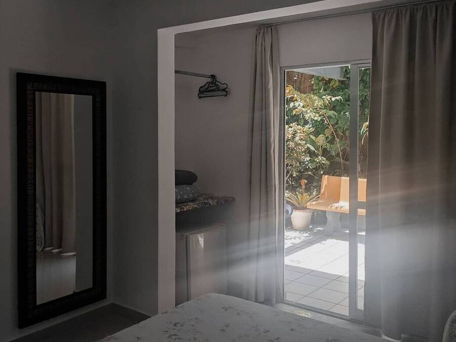 Een slaapkamer met een bed, een spiegel, een klein tafeltje en uitzicht op een buitenruimte met planten en een bankje.