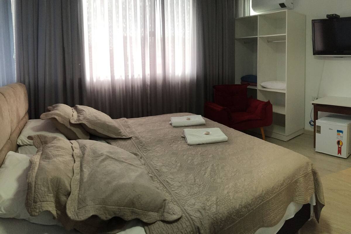 A neatly made bed with a beige blanket, a red chair, a TV mounted on the wall, and a small table with a refrigerator.
