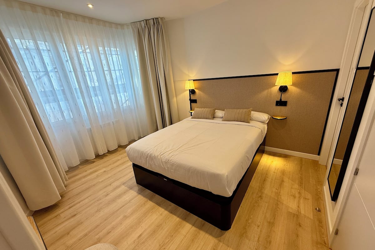 A hotel room with a large bed, wooden flooring, a headboard with wall-mounted lights, and sheer curtains.