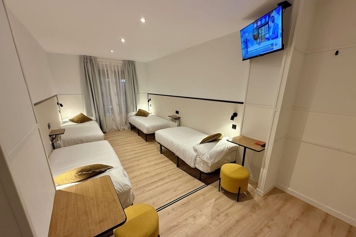 A hotel room with three single beds, a wooden table, a yellow ottoman, a flat-screen TV mounted on the wall, and a window with curtains.