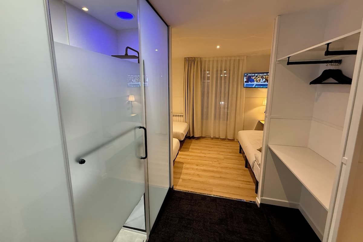 A hotel room with a glass partition, a bed, a TV, a wardrobe, and a shower area.