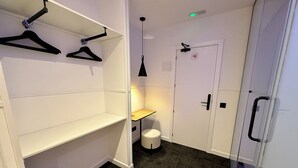 A compact room with white walls, a small wooden table, a white door with a handle, and a black hanger on a white shelf.