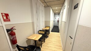 A corridor with wooden flooring, black chairs, and small wooden tables. A fire extinguisher is mounted on the wall.