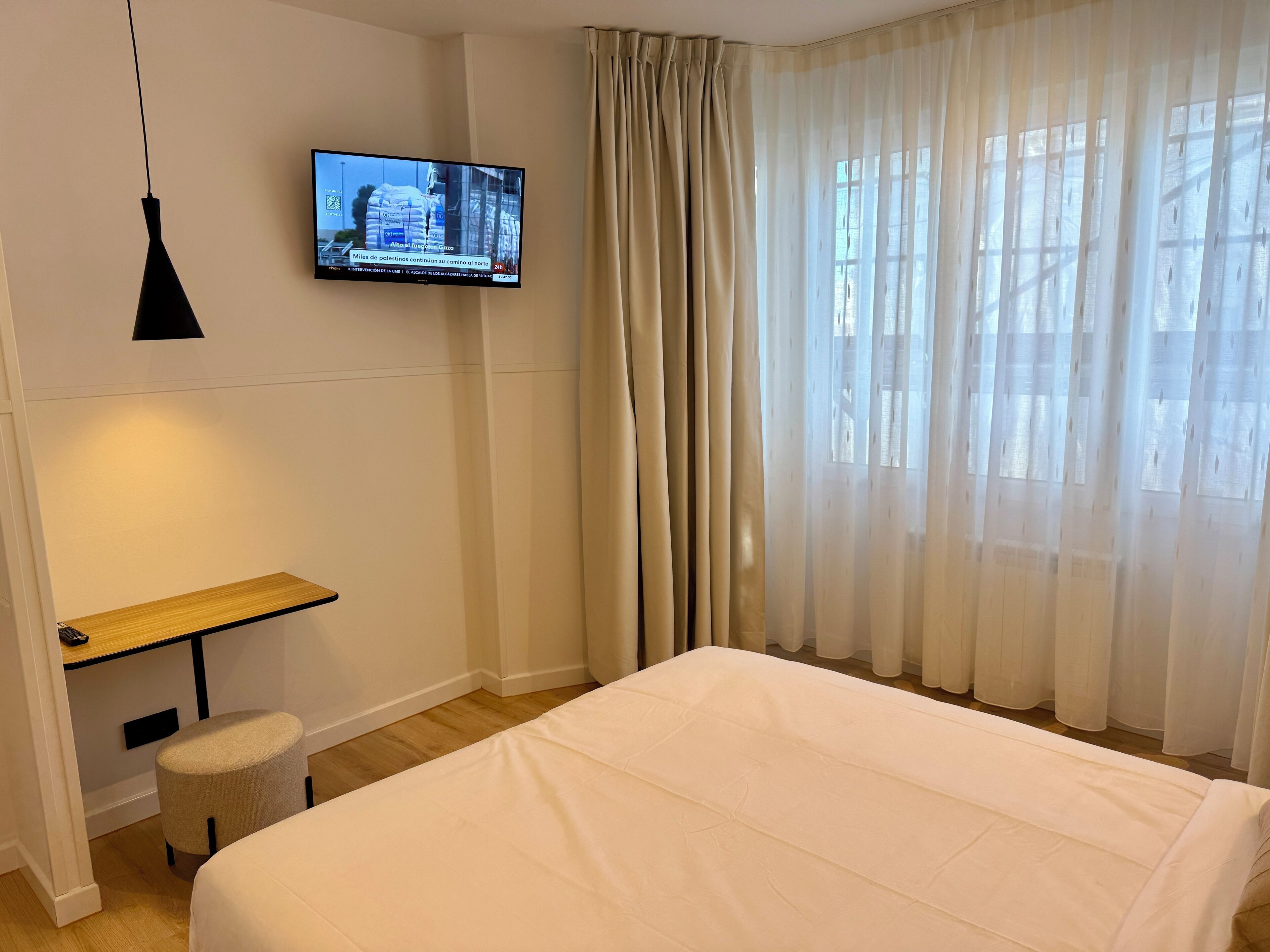 Et hotelværelse med seng, skrivebord, stol, fjernsyn ophængt på væggen og et vindue med lette gardiner.