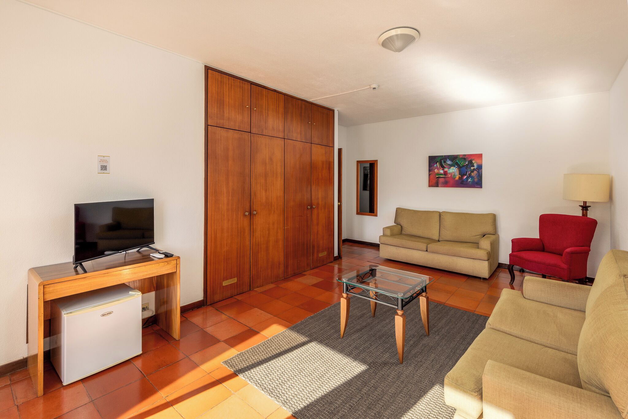 Uma sala de estar com televisão, sofá, poltrona vermelha, mesa de centro de vidro e armário de madeira.