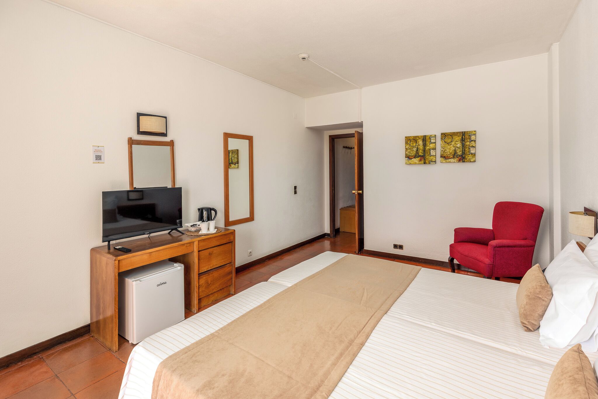 A hotel room with a bed, a desk with a television, a small refrigerator, a red chair, and artwork on the wall.