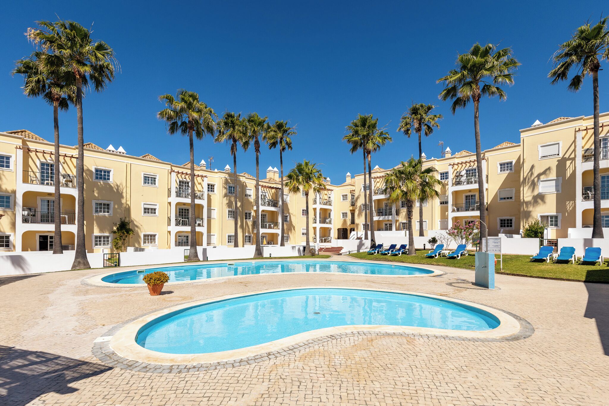 A pool area with palm trees, lounge chairs, and a building with balconies.