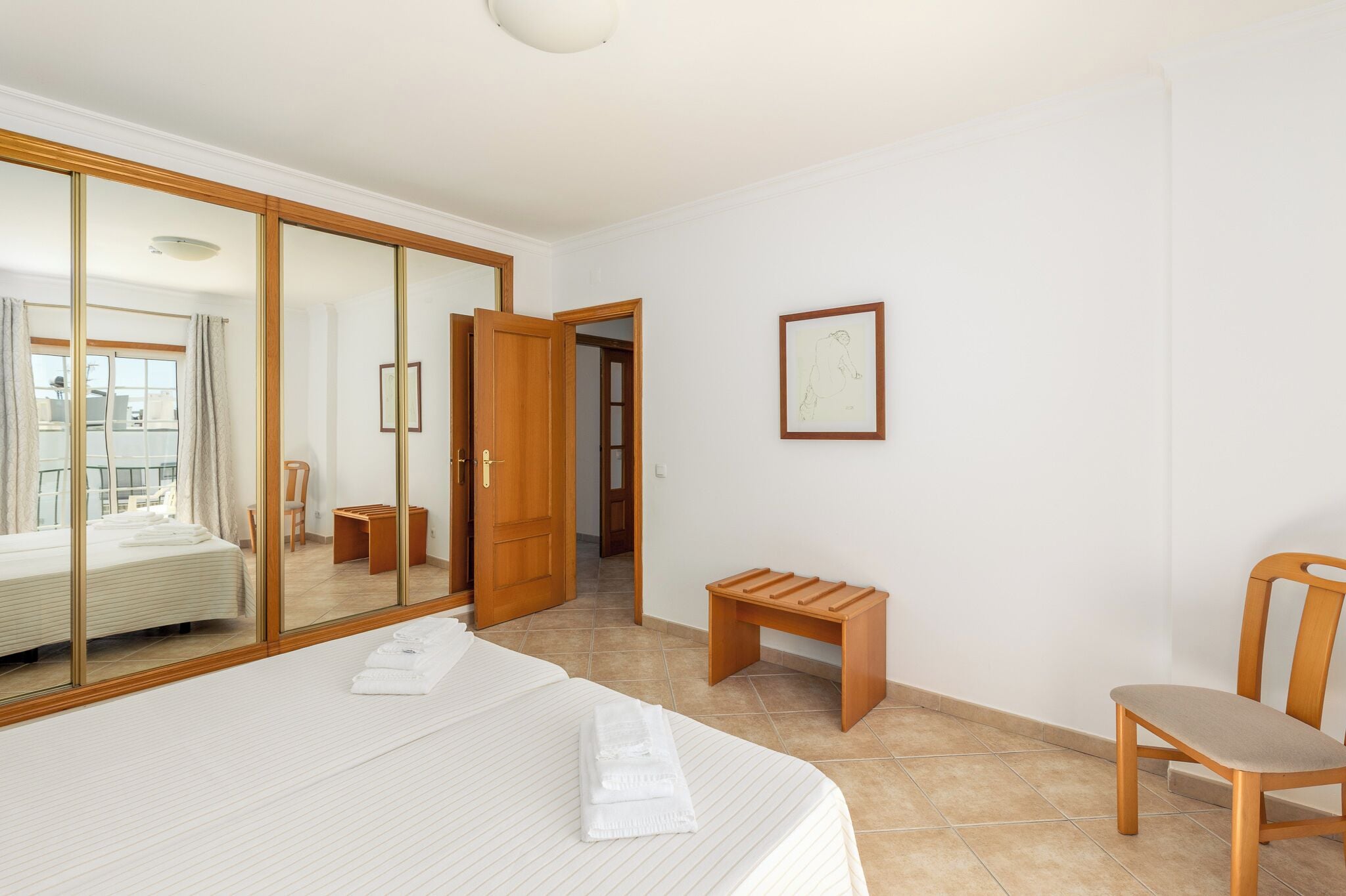 Una habitació d'hotel amb llit, una banca de fusta, una cadira, un mirall i una porta que dona a una altra habitació.