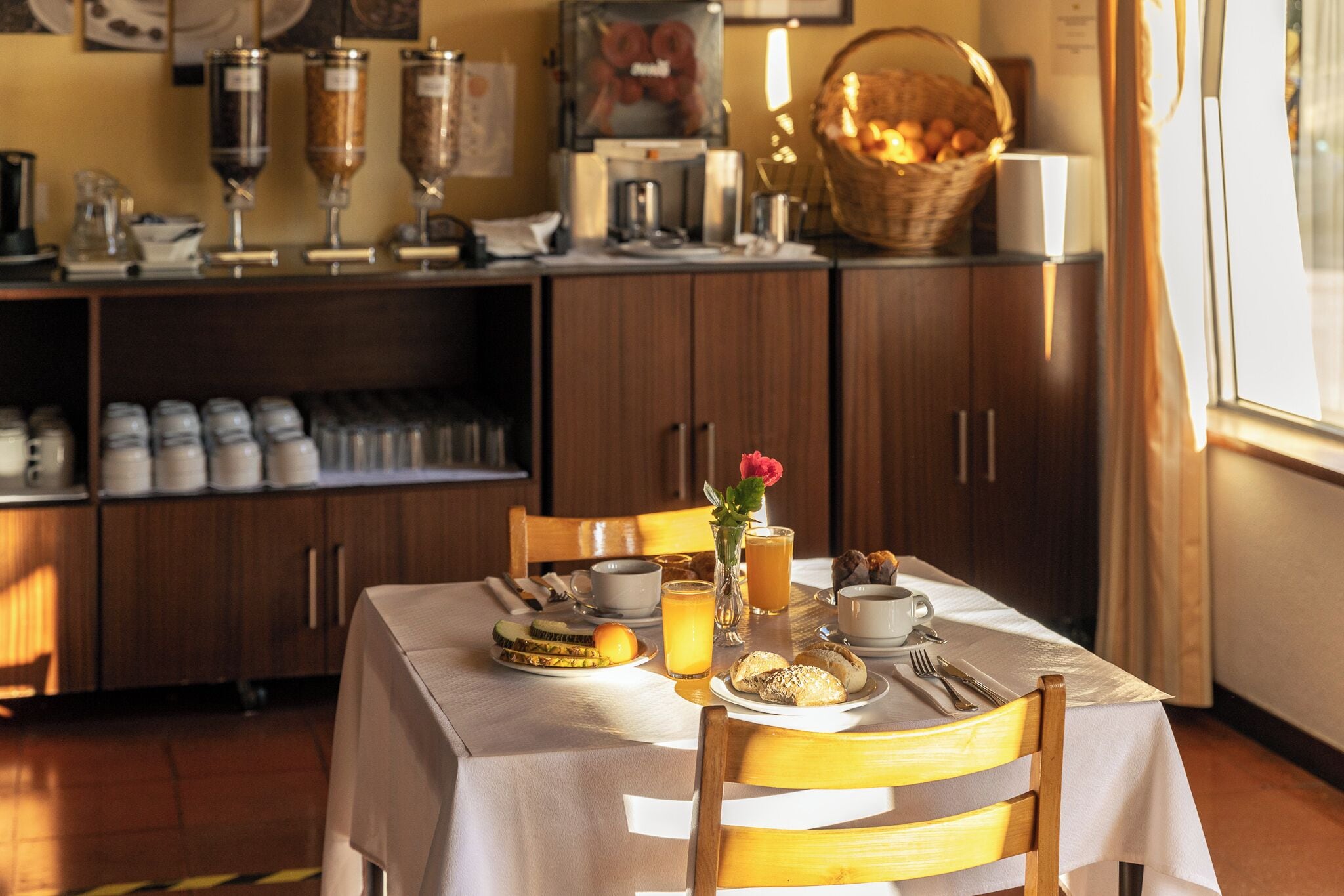 A breakfast table set with utensils, a vase with a flower, and a basket of bread.