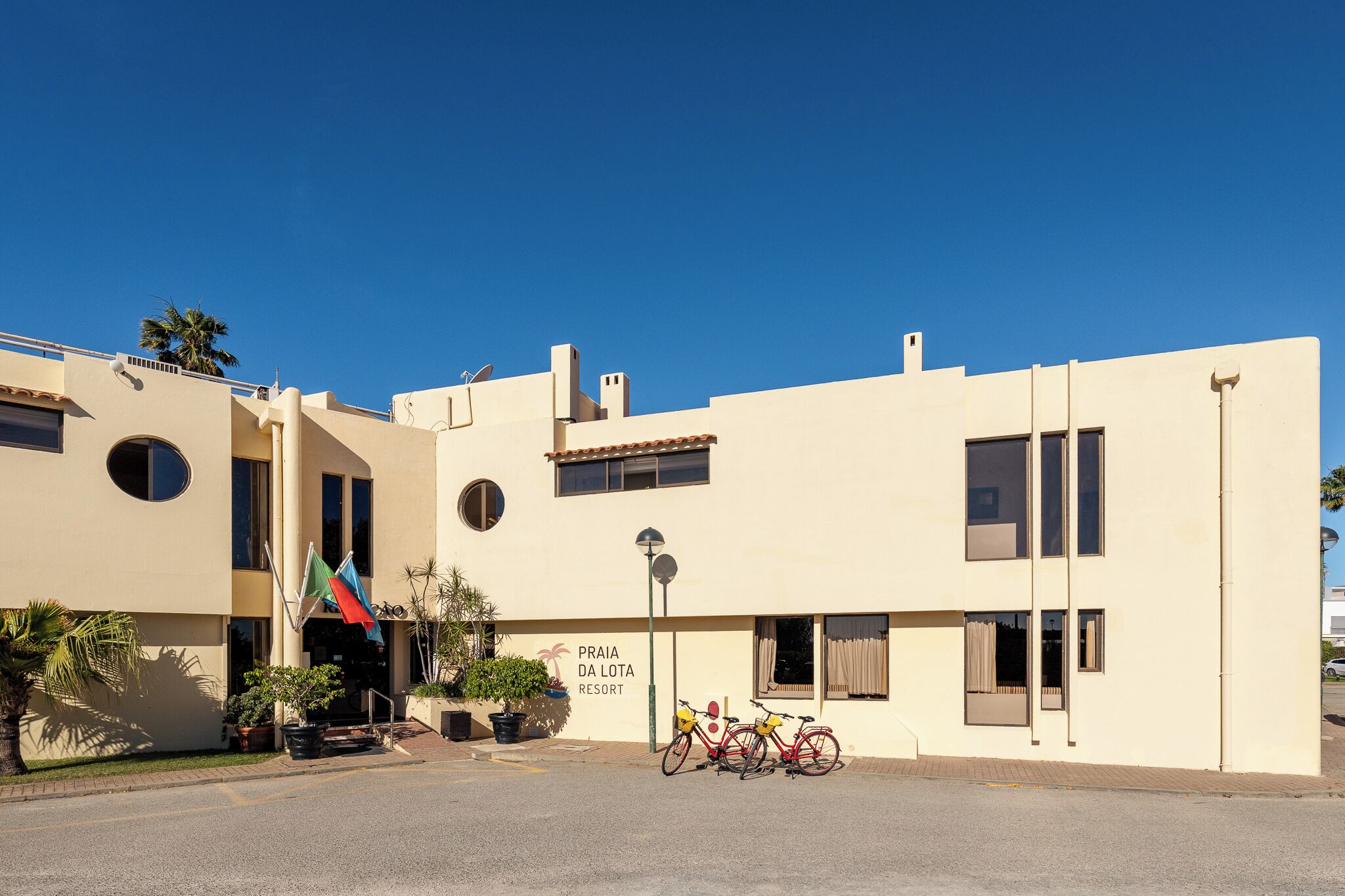 Een gebouw met een bord waarop 'Praia da Lota Resort' staat, met een moderne architectuur, een plat dak, grote ramen en een verharde entreezone met fietsen ervoor.