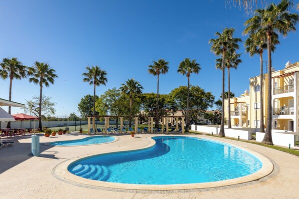 A swimming pool with palm trees, a covered seating area, and a building with balconies.