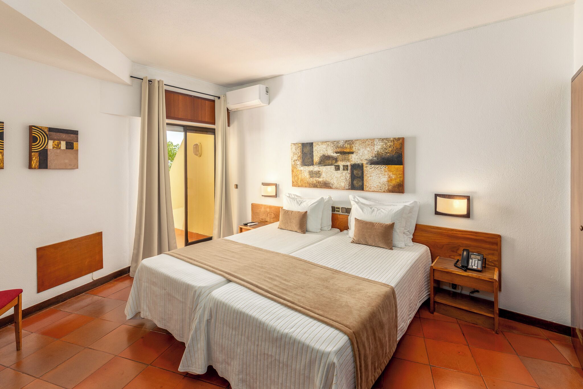 Ein Hotelzimmer mit zwei Betten, einem Holz-Kopfteil, einem Nachttisch mit Telefon und einem Fenster mit Vorhängen.