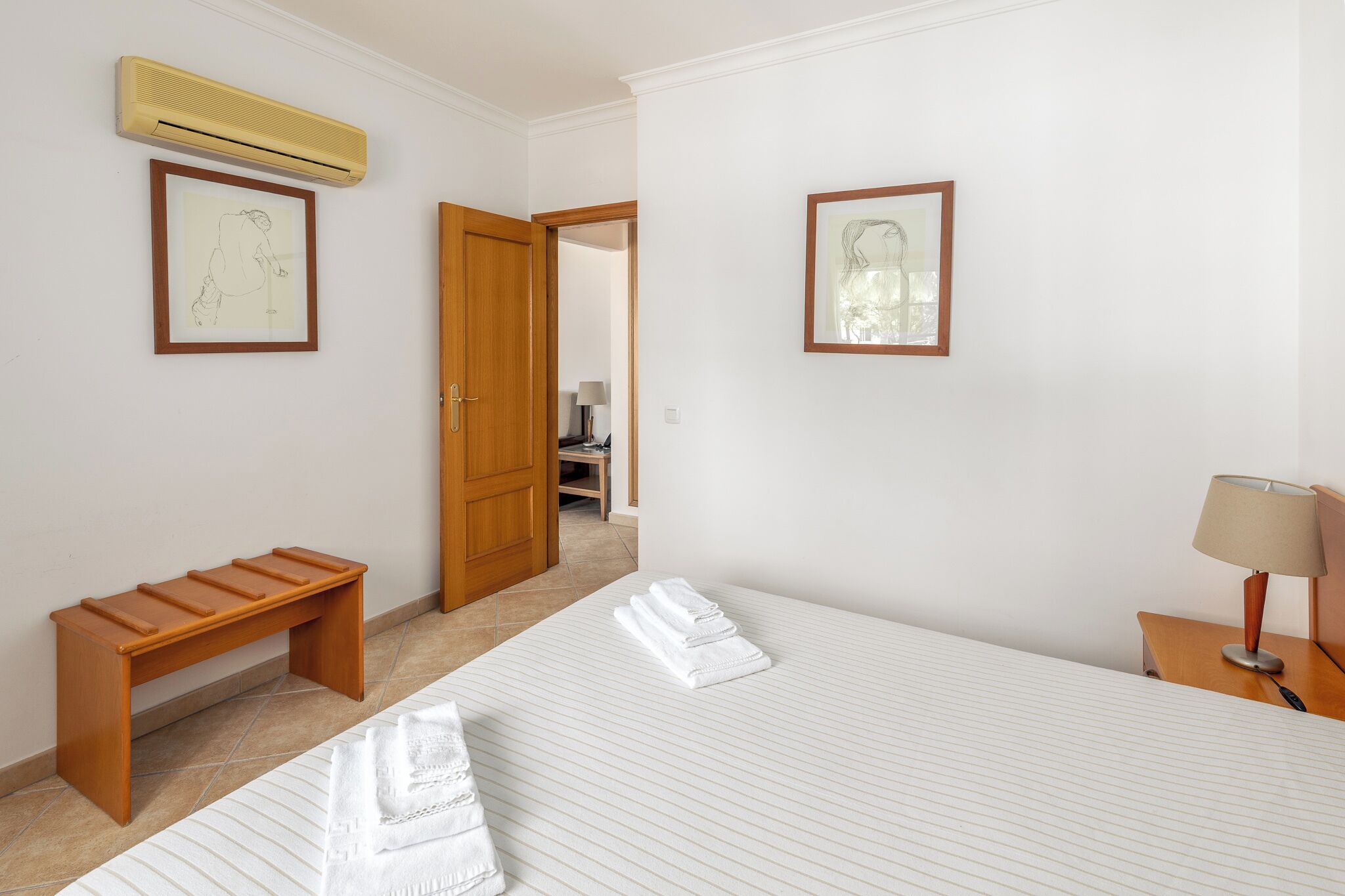 A hotel room with a bed, a wooden bench, an air conditioner, and two framed pictures on the wall.
