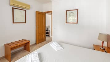 A hotel room with a bed, a wooden bench, an air conditioner, and two framed pictures on the wall.