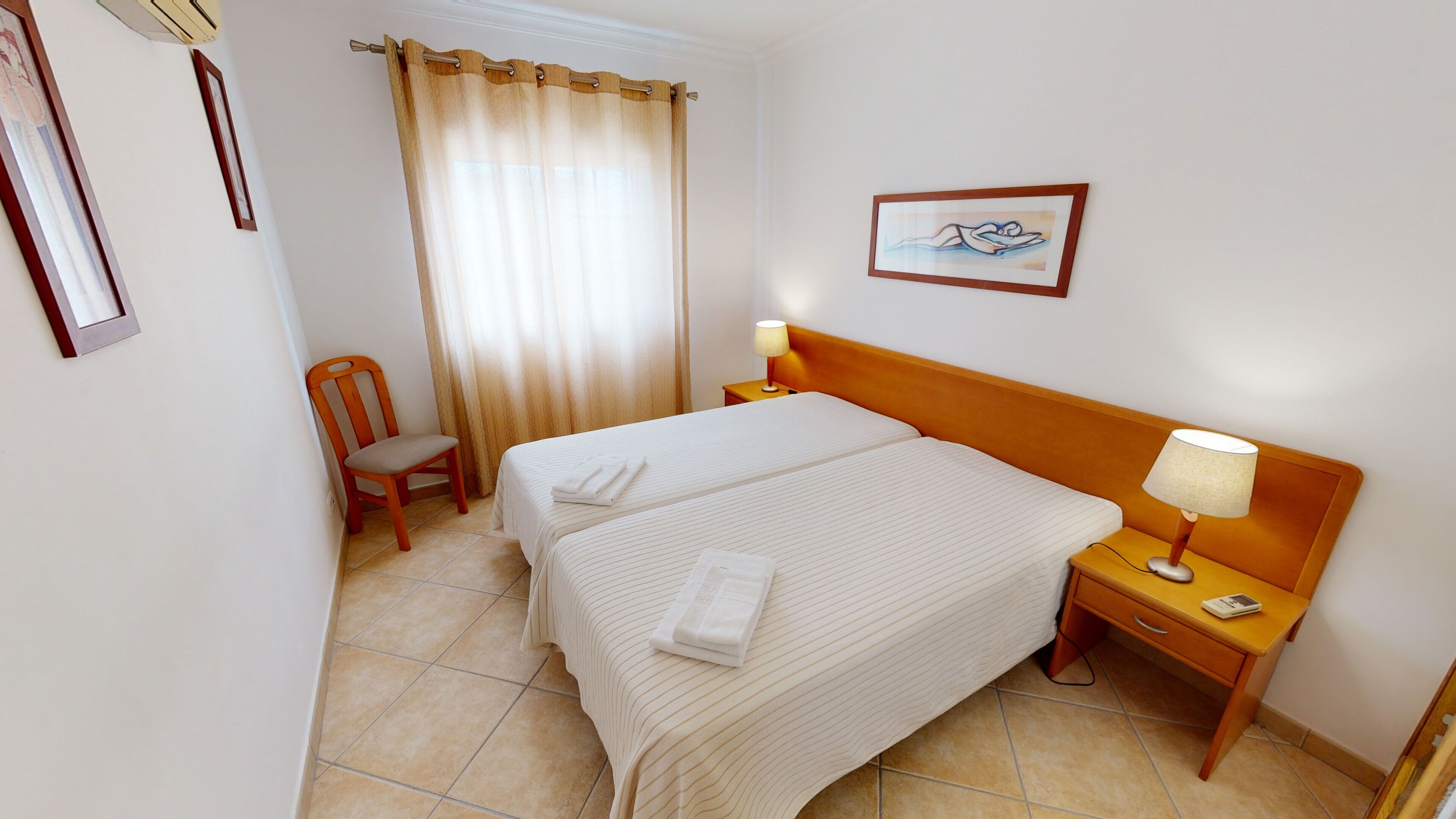 Habitación de hotel con dos camas, una silla, una mesita de noche con lámpara y un cuadro en la pared.