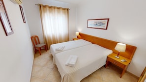A hotel room with two beds, a chair, a nightstand with a lamp, and a framed picture on the wall.