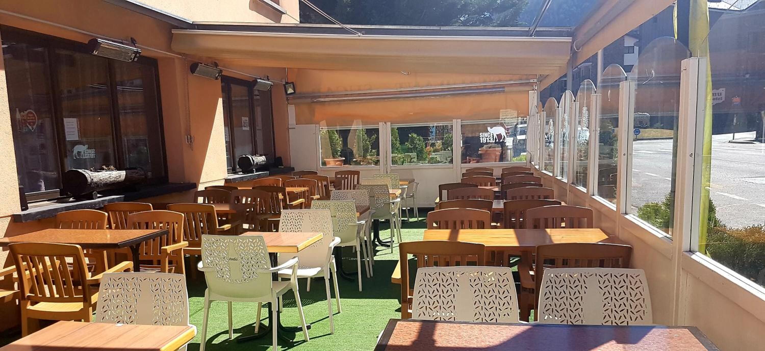 An outdoor café with wooden tables and chairs, a green artificial turf floor, and large windows offering views of the street.