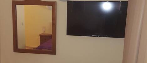 Una habitación pequeña con una mesa y sillas de madera, un espejo, un televisor montado en la pared y un cubo de basura.
